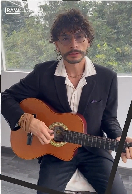 A person wearing a black suit and glasses holding a guitar, sitting in a well-lit room with greenery visible through a window.