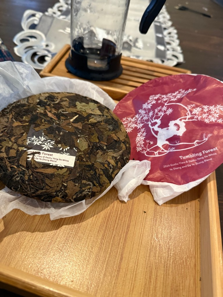 A close-up of a tea cake and its packaging on a wooden tray, with a glass tea infuser in the background.