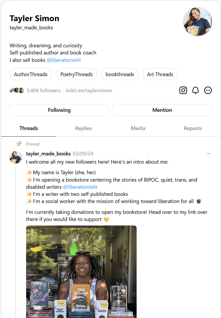 A smiling individual stands in front of a window display of books, holding two books. The display features titles by various authors, emphasizing a community-focused bookstore.