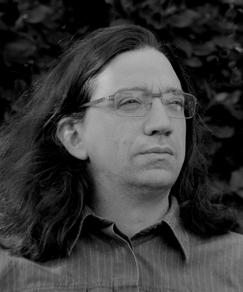 A black and white portrait of a man with long hair and glasses, looking thoughtfully into the distance.