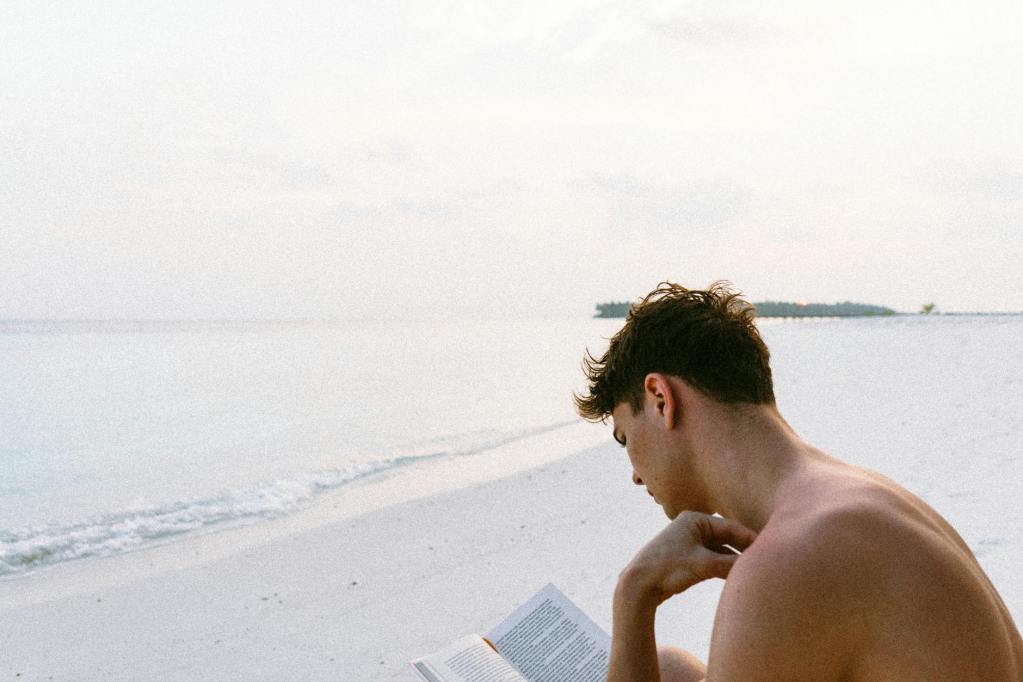 A young man sitting on a beach reading a book, with a calm sea and gentle waves in the background.