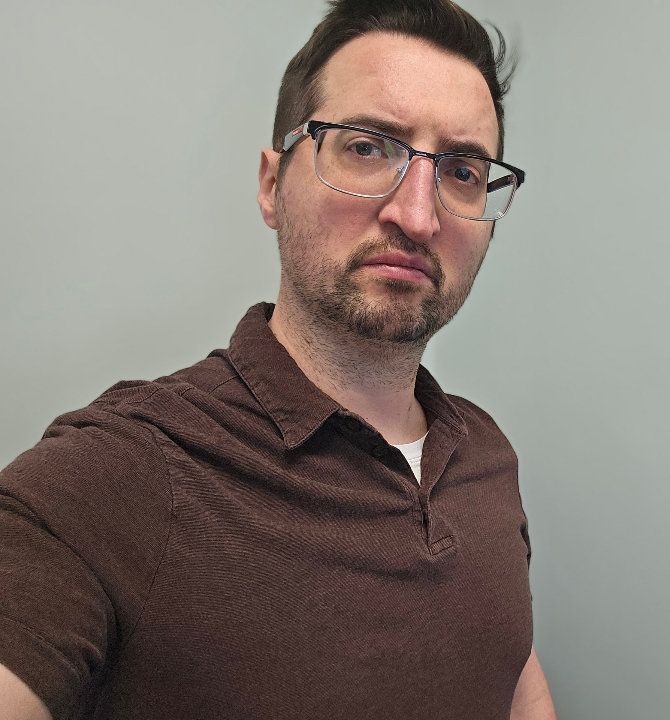 A close-up selfie of a person with glasses and medium length hair, wearing a brown polo shirt, with a neutral expression, set against a light gray background.